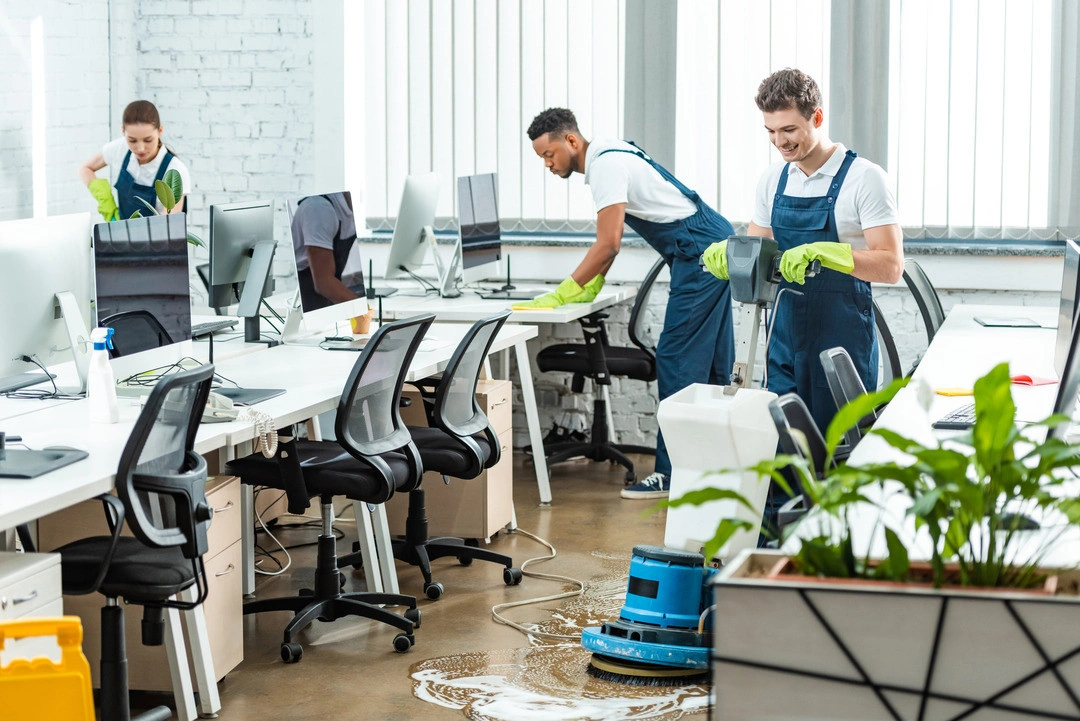 Two cleaners in overalls and gloves clean an office space with computers, chairs, and potted plants, while a blue machine is in use.