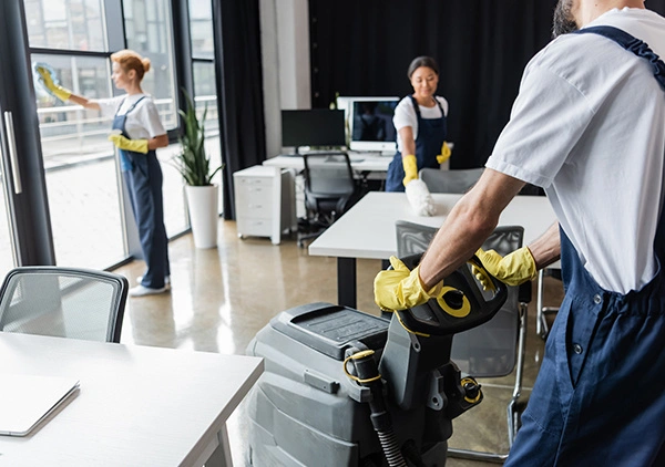 A team of cleaners in a modern office setting, using various cleaning tools, with neatly arranged furniture and large windows.